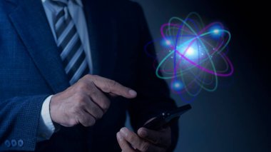 İş adamı, karanlık arka planda soyut parlayan atom hologramı olan akıllı telefon kullanıyor. Nükleer füzyon konsepti, sonsuz enerji, ucuz elektrik, gelecek teknolojileri. Bilim ve gelecek kavramı