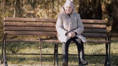 Gri kareli ceketli ve gri bereli yaşlı bir kadın bir bankta oturuyor. Şeffaf bir şemsiye ayaklarının yanında duruyor. Bankın üzerine altın sonbahar yaprakları serpti ve onları bir araya topluyor.