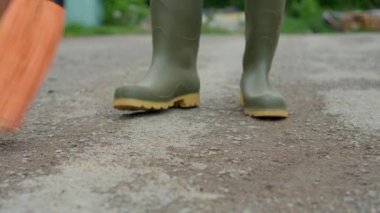 Yeşil lastik çizmeli biri, çakıllı yolda süpürgeyle toprağı süpürür, temizlik ve peyzaj sergiler..