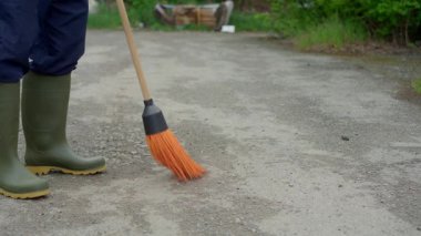 Yeşil çizmeli bir adam, turuncu süpürgeyle etrafı dikkatlice süpürüyor.