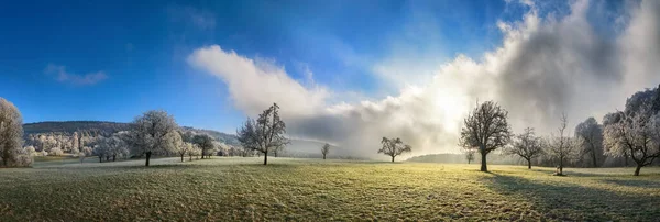 Winter landscape panorama, hoarfrost on open meadow, blue sky with dramatic clouds and sun rays