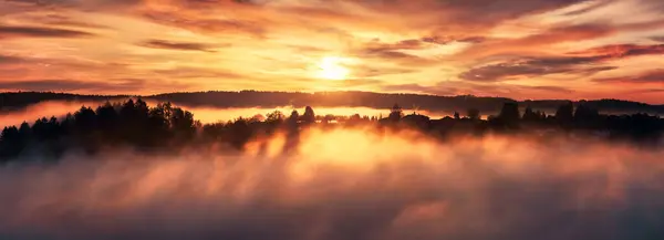 雾气笼罩下的日出 山上一幅幅树木的轮廓 梦幻般的天空 升起的太阳和温暖的色彩 尽收眼底 图库照片