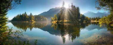 Suya yansıyan dağlar ve ağaçlarla resmedilmiş göl manzarası. Güzel güneş ışınları olan, cennet gibi panoramik bir manzara.