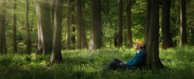 Ormandaki ağaçların altında oturan genç adam, güneşin sakin ışığına bakıyor. Panoramik biçim.