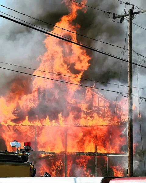 Photo of raging house fire with flames and thick gray and black smoke rising from it. Part of a fire truck can be seen in the foreground with a red light. Smoke curls around a telephone pole. Electrical wires are getting scorched. Smokey scene.