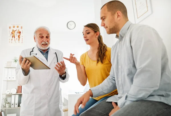 Senior doctor holding tablet talking to young couple in his office in clinic
