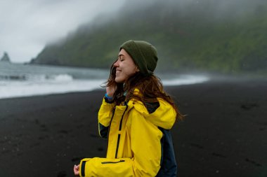 Sarı ceketli bir kız sisli bir sahil şeridinde, siyah kumlu bir arka planda duruyor..