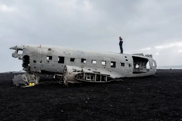 Terk edilmiş bir uçak kazası ve üstünde siyah bir volkanik plajda duran bir adam.