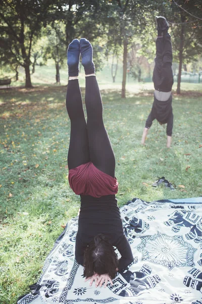 年轻女子在户外公园做瑜伽姿势 男子背景做手倒立 — 图库照片