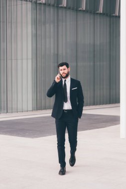 Young bearded elegant businessman walking outdoors talking smartphone