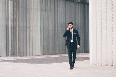 Young bearded elegant businessman walking outdoors talking smartphone
