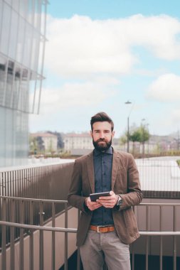 Young bearded man outdoors posing looking camera holding tablet computer smiling serene and confident