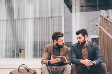 Two elegant contemporary bearded businessman outdoors sitting staircase sharing tablet remote working discussing brainstorming