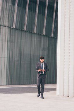 Young bearded elegant businessman walking outdoors using smartphone