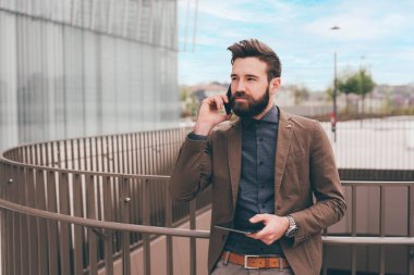 Genç, sakallı, modern iş adamı dışarıda elinde tabletle akıllı telefonlardan konuşuyor.