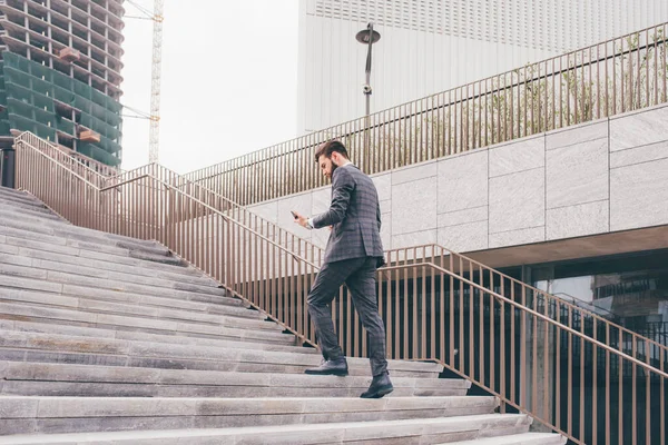 若い髭のエレガントなビジネスマンが2階に行く屋外でスマートフォンを使用して歩く — ストック写真