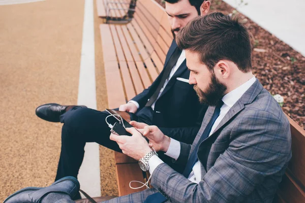 スマートフォンを使用して市内の屋外のベンチに座っている2人の若い現代的なビジネスマン ビジネス コミュニケーションコンセプト — ストック写真