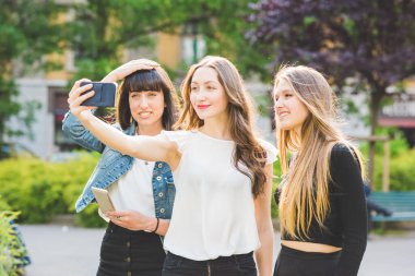 Üç güzel beyaz kadın, milenyum çağında, akıllı telefon ile selfie çekiyorlar. Şehirde açık havada, gülümsüyorlar. Sosyal ağ, teknoloji, arkadaşlık kavramı.