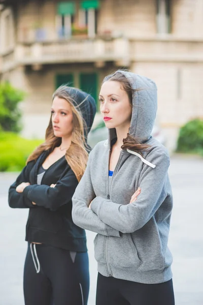 Two young millennial sportive woman wearing tracksuit outdoor in the city, arms crossed.