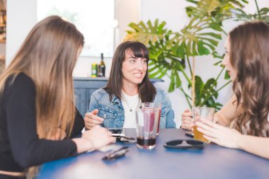 Üç genç kadın barın içinde oturmuş sohbet ediyor ve içki içiyor.