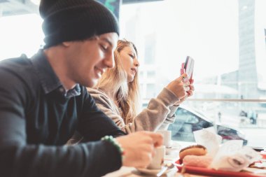 İki genç arkadaş akıllı telefon kullanarak kahvaltı yapıyor.