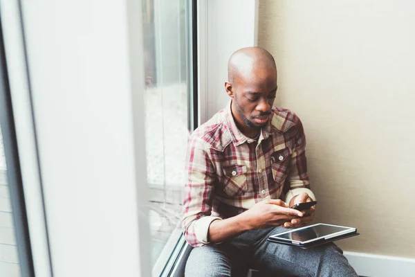 Hombre Negro Sentado Cerca Ventana Interactuando Con Teléfono Inteligente — Foto de Stock