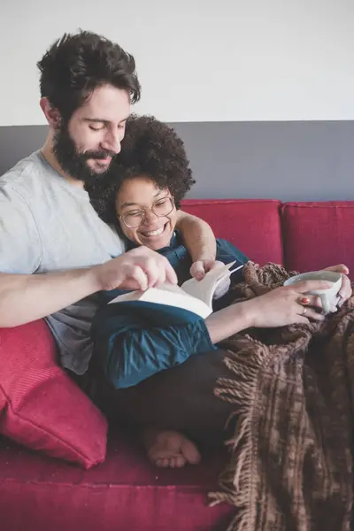 Young Multiethnic Couple Hugging Couch Reading Book — Stock Photo, Image