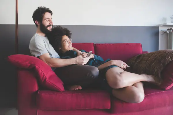Pasangan Muda Multietnis Yang Memeluk Sofa Dalam Rumah Menggunakan Smartphone — Stok Foto