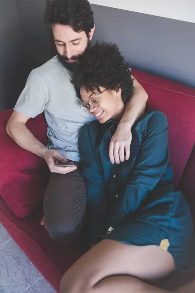 Young Multiethnic Couple Using Smart Phone Indoor Sitting Couch Hugging — Stock Photo, Image