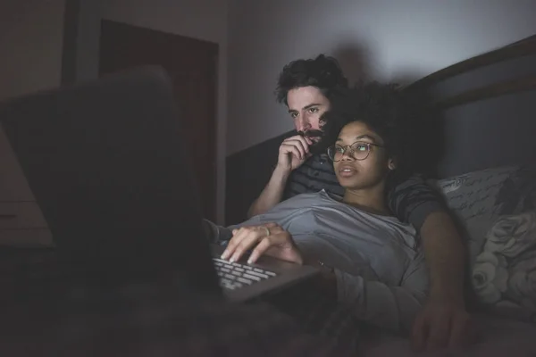 Pasangan Muda Multietnis Dalam Ruangan Menggunakan Komputer Duduk Tempat Tidur — Stok Foto