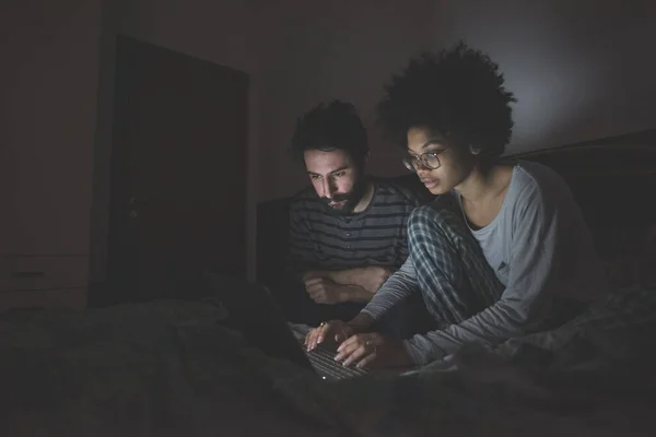 Joven Pareja Multiétnica Interior Usando Computadora Acostado Cama Por Noche Fotos de stock libres de derechos