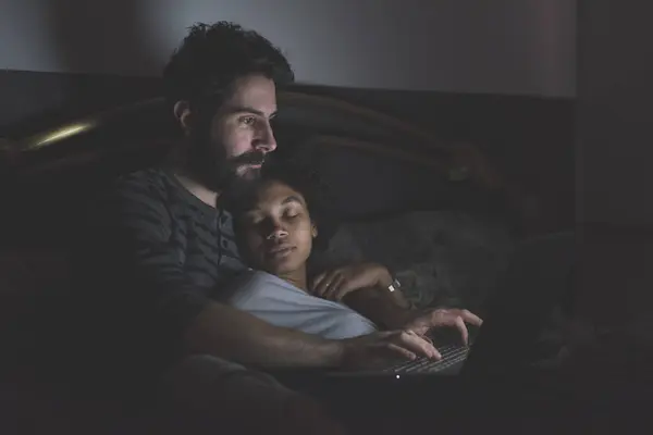 若い多民族のカップル 夜にコンピュータの横たわるベッドを使用して屋内 ロイヤリティフリーのストック画像