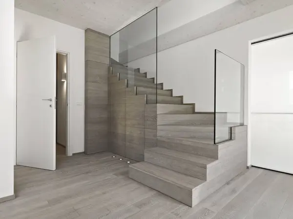 Close-up of a modern wooden staircase with a glass railing in the living room. The wooden floor is visible on the left, and there is an open white door.