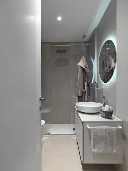右の前景にモダンなバスルームインテリア カウンタートップ洗面台付き洗面台キャビネットとその上に壁にぶら下がる丸い鏡 背景にはガラスドア付きのメイソンシャワーキュービクルがあります — ストック写真