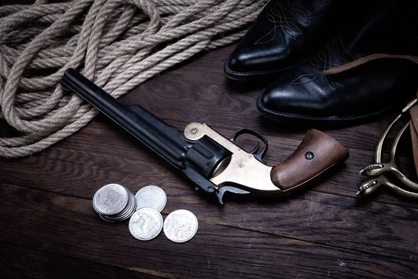 Old west revolver gun with silver dollars with hat, rope and cowboy boots on wooden table background.