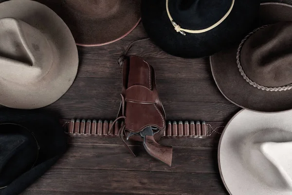 Gun with holster and gun belt with old west hats on wooden table. Top view.