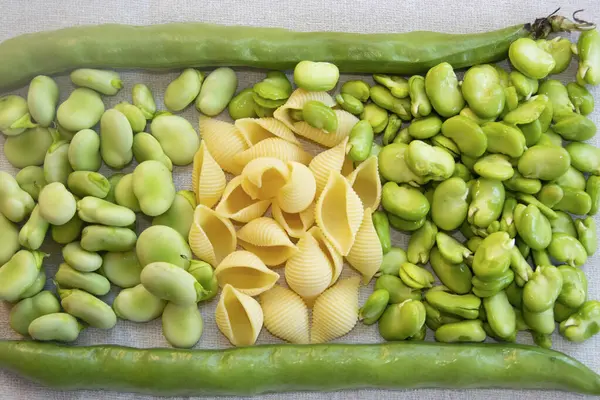 Keten kumaş üzerinde taze fasulye ve makarna kabukları. Canlı yeşil fasulye ile soluk sarı makarna. Rustik besin bileşimi, sağlıklı malzemeler, Akdeniz mutfağı, İtalyan yemekleri, organik sebzeler, bahar hasadı.