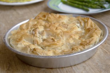 Yakın plan: Köy ahşap bir masada yuvarlak bir alüminyum tavada taze pişmiş, altın sarısı filo pastası. Çıtır çıtır, pul pul parlıyor. Arka planda hafif bulanık mezeler.