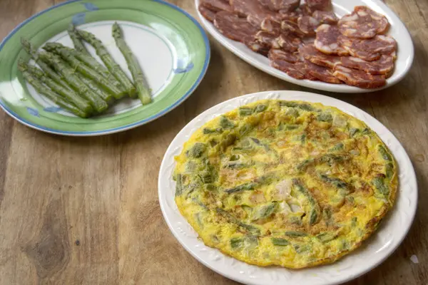 Kırsal bir masada beyaz bir tabakta taze yeşil kuşkonmaz ile altın omlet. Arka planda görünen beyaz kuşkonmaz