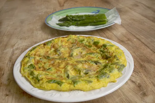 Yeşil kuşkonmaz ve parmesanlı ev yapımı omlet, kırsal ahşap bir masada beyaz bir tabakta. Arka planda beyaz kuşkonmaz. Sağlıklı kahvaltı ve kahvaltı için mükemmel..