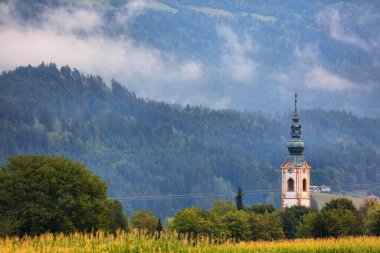 Gailtal Alplerindeki sisli orman katmanları. Avusturya 'nın Carinthia bölgesinde yağmur üstüne kilise kulesiyle dağ manzarası.