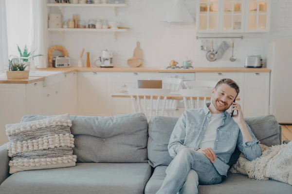 stock image Happy smiling young man with stubble in casual clothes talking on phone with his friend while sitting with crossed legs on cozy couch, kitchen counter in blurred background. Communication concept