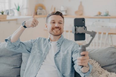 Videodaki genç adam memnuniyetini ifade ediyor, elinde telefonla el sallıyor, başparmak hareketi yapıyor ve koltukta otururken kameraya gösteriyor, geçmişi bulanık.