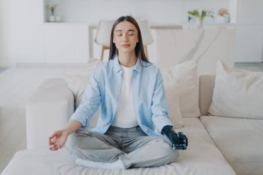Sakin, engelli bir kız yoga yapıyor, meditasyon yaparken nilüfer pozisyonunda oturuyor. Genç dişi, evdeki kanepede biyonik protez koluyla mudra hareketi yapıyor. Özürlü insanların sağlıklı yaşam tarzı.