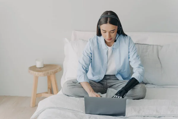 Female student with disability wearing headset studying online at laptop, typing, using bionic prosthetic arm, sitting on bed at home. Elearning, distant job of disabled people concept.