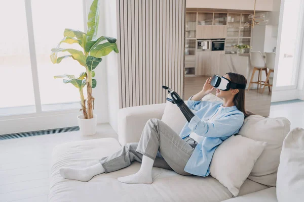 Disabled girl with artificial limb wearing virtual reality glasses learning to use bionic prosthetic arm during rehabilitation after limb loss, sitting on sofa in cozy living room. Medical high tech.