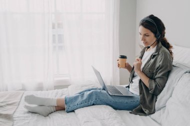 Kulaklık takan bayan koçun dizüstü bilgisayara bakışı web kameralı bir konferansa ev telefonundan bağlanıyor. Kulaklıklı kadın yabancı dil öğretmeni bilgisayar başında, yatakta oturarak ders veriyor..