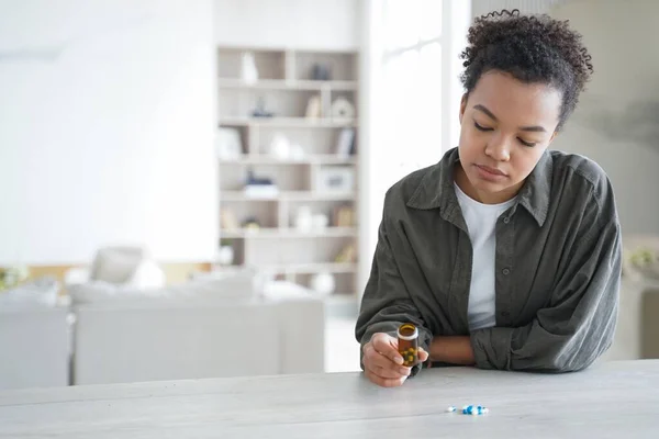 Doubting young mixed race girl holding jar with medicines before taking meds. Pensive female holds medicines doubts whether to take medical pills medications, sitting at table at home.