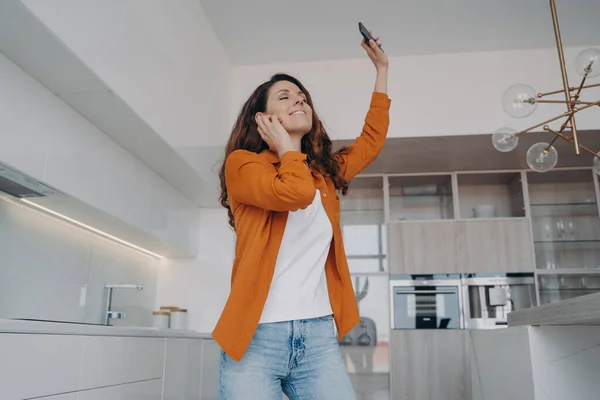 Happy woman holding smartphone, dancing alone in kitchen at home. Smiling female with closed eyes dances listening to favorite music on phone, moving to song, enjoying musical sound.