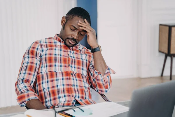 Tired african american businessman suffering from headache, after working at laptop, sitting at workplace. Overworked black male guy touching head, pondering problem. Stress at work concept.